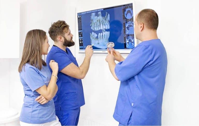 Trzech dentystów w niebieskich fartuchach bada zdjęcie rentgenowskie zęba na ekranie. Jeden z nich trzyma model dentystyczny, podczas gdy drugi wskazuje na zdjęcie rentgenowskie, podkreślając Profesjonalny Stomatolog Kraków podczas ich szczegółowej dyskusji.