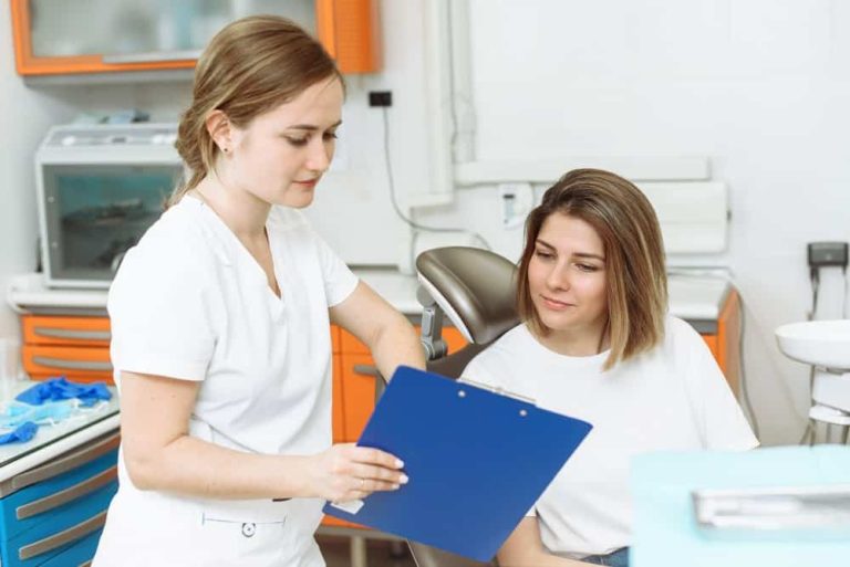 Profesjonalny dentysta Kraków w białym fartuchu pokazuje niebieski schowek siedzącej pacjentce w nowoczesnej klinice dentystycznej z pomarańczowymi szafkami w tle.