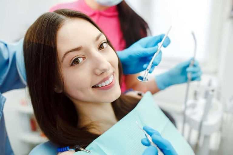 Młoda kobieta siedzi na fotelu dentystycznym, uśmiechając się do kamery, podczas gdy Profesjonalny Stomatolog Kraków w rękawiczkach trzyma narzędzia dentystyczne w pobliżu jej ust. W tle stoi inny członek personelu dentystycznego.