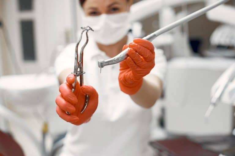 A Profesjonalny dentysta Kraków w czerwonych rękawiczkach i maseczce prezentuje narzędzia, takie jak kleszcze do ekstrakcji i wiertło, w jasno oświetlonym gabinecie.